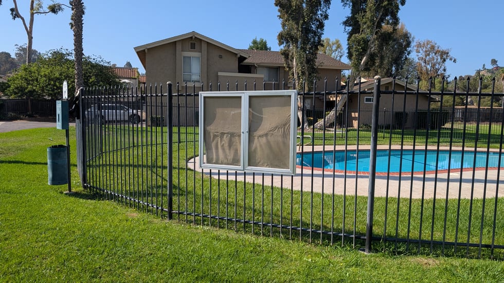 A metal, glass-fronted, two-door lockable notice case mounted on the fence beside the pool on the Amistad Place side. Both panes are backed with plain paper and display no notice.