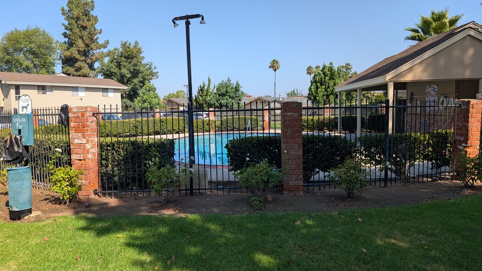 The pool enclosure on the Amistad Court side of Jamacha Greens, seen from outside the black perimeter fence: the pool, clipped hedges, brick pillars, a pet waste station and a litter bin at the left, pool signage on the far fence. No notice board is mounted anywhere in view.