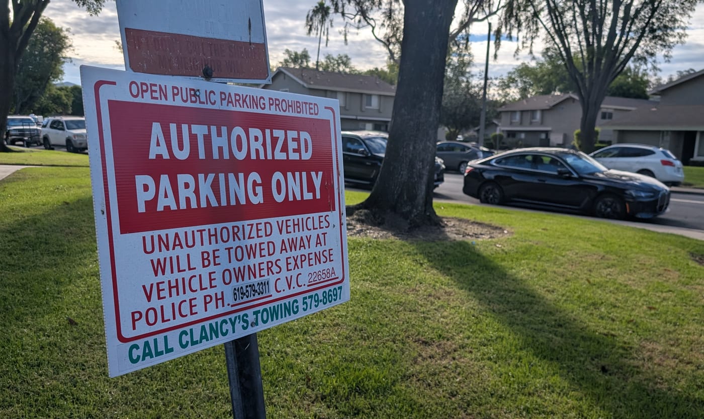 A parking sign on a steel post in the lawn at the edge of the private lot off Gustavo Street. It reads OPEN PUBLIC PARKING PROHIBITED across the top, then AUTHORIZED PARKING ONLY in white on a red panel, then UNAUTHORIZED VEHICLES WILL BE TOWED AWAY AT VEHICLE OWNERS EXPENSE, POLICE PH. 619-579-3311, C.V.C. 22658A, and CALL CLANCY'S TOWING 579-8697 in green along the foot. An older sign on the same post above it has weathered until only IF YOU DON'T CALL THE SHERIFF MY NEIGHBOR WILL can still be read. Cars are parked in the lot behind the sign and along the public street beyond it, with a mature tree and two-storey units behind them.